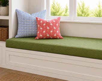May include: A white built-in window bench with a solid olive green cushion. Two square throw pillows are placed on the bench: one features a blue and white grid pattern, and the other is a coral pink pillow decorated with a repeating white flamingo print.