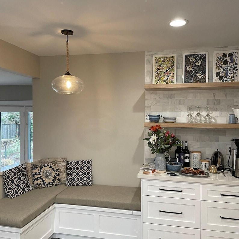 Modern kitchen nook with custom L-shaped bench cushions in taupe fabric, white cabinetry, and open wood shelving. Accented with patterned pillows, marble backsplash, pendant light, and fresh flowers