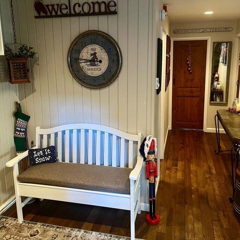 Cozy holiday entryway with white bench featuring a custom brown cushion, nutcracker decor, and a ‘Let it Snow’ pillow. Wood floors, wall clock, and welcome sign add warm farmhouse charm
