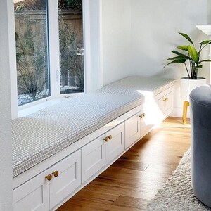 Bright window seat with custom gray patterned bench cushion on white built-in drawers with brass knobs. Modern cozy reading nook with natural light, hardwood floors, and a potted plant accent