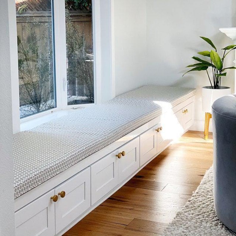 Bright window seat with custom gray patterned bench cushion on white built-in drawers with brass knobs. Modern cozy reading nook with natural light, hardwood floors, and a potted plant accent