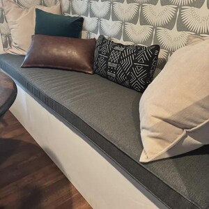 Modern dining nook with custom dark grey bench cushion and mix of leather, geometric, and neutral throw pillows. Patterned wallpaper adds contrast for a cozy, contemporary home seating area