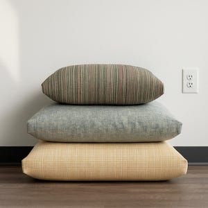 May include: Three decorative pillows stacked on top of each other. The top pillow has vertical stripes in shades of green, brown, and red. The middle pillow is a solid light blue-gray. The bottom pillow is a light yellow with a woven texture.