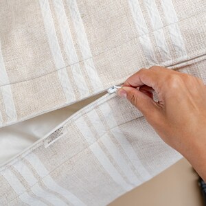 May include: Close-up of a beige and white striped pillow cover with a zipper closure. The pillow cover has a textured fabric and a white zipper. A hand is pulling the zipper open.