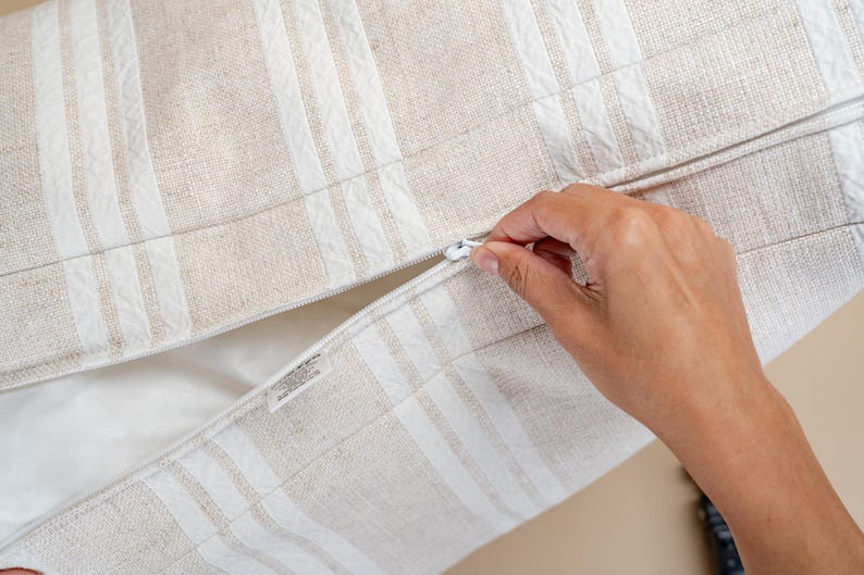 May include: Close-up of a beige and white striped pillow cover with a zipper closure. The pillow cover has a textured fabric and a white zipper. A hand is pulling the zipper open.