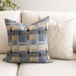 May include: A square throw pillow featuring a blue, tan, and black plaid pattern with a visible woven texture, positioned next to a second square throw pillow in a solid cream color with an intricate, tone-on-tone embroidered leaf pattern.