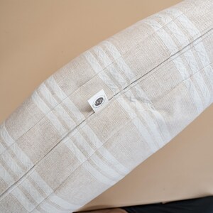 May include: A rectangular pillow with a beige and white plaid pattern. The pillow has a zipper closure and a small tag with text. The pillow is made of a soft, textured fabric.