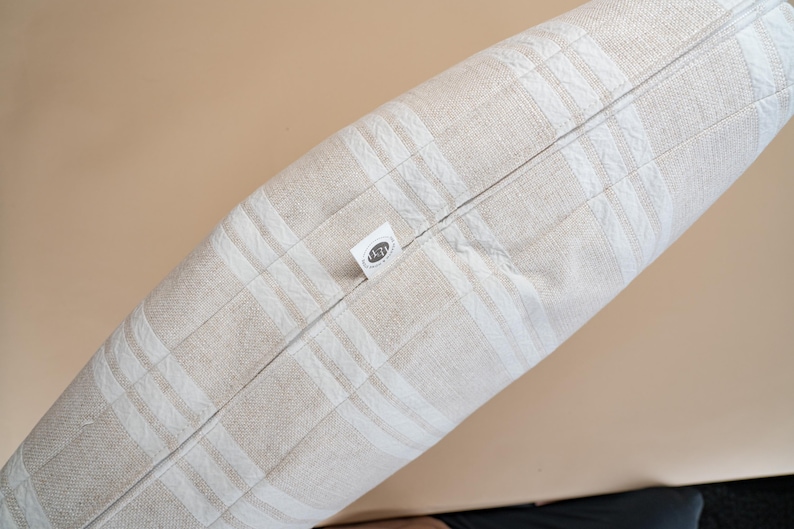May include: A rectangular pillow with a beige and white plaid pattern. The pillow has a zipper closure and a small tag with text. The pillow is made of a soft, textured fabric.