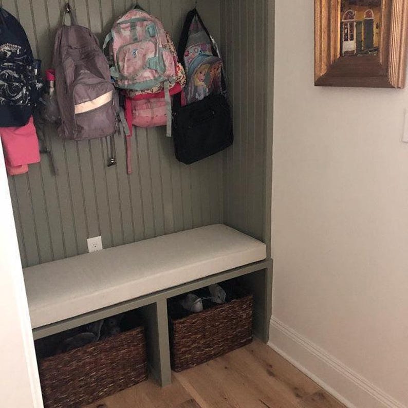 Custom mudroom bench cushion in light neutral fabric with beadboard paneling, woven storage baskets, and kids’ backpacks hanging above. A functional and cozy entryway seating solution.