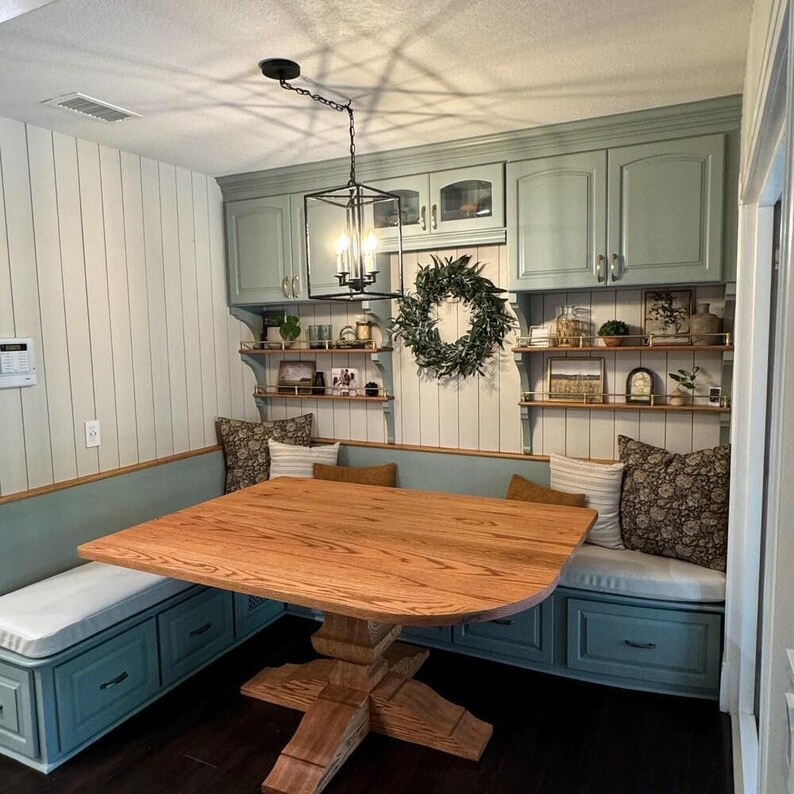 Cozy farmhouse breakfast nook with custom bench cushions in light fabric, sage green built-in seating, wood table, and decorative pillows. Features cabinets, open shelving, and a wreath accent for warm cottage charm.