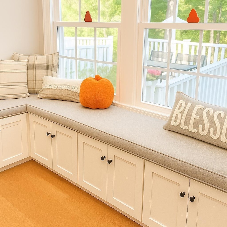 Cozy fall window seat with custom neutral bench cushion, plaid and fringe pillows, knit orange pumpkin, and ‘Blessed’ pillow. White built-in seating with cabinets adds farmhouse charm and autumn decor.