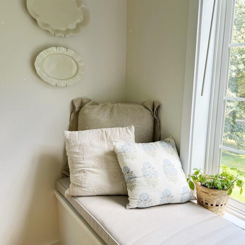 Cozy window seat with neutral linen cushion and embroidered pillows in cream and soft blue. Features a simple farmhouse look with ceramic wall plates and a small potted plant by the window