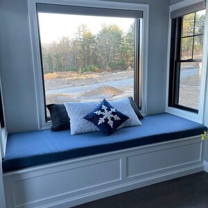 Cozy window seat with custom blue bench cushion and winter-themed throw pillows, including a snowflake design. Large windows bring in natural light and a view of the outdoors for a peaceful nook setting.