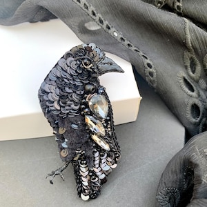May include: A black crow brooch made of sequins and rhinestones. The crow is facing to the right and has a detailed design.