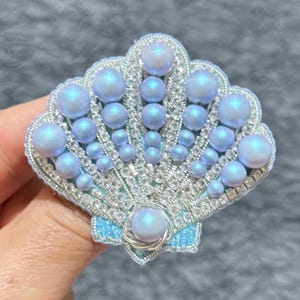 以下が含まれることがあります： 淡い青と銀色のビーズでできた貝殻のブローチ。虹色の真珠が飾られています。