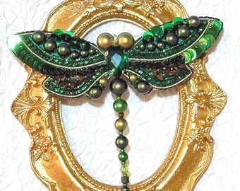May include: A green dragonfly brooch made of sequins, beads, and pearls. The dragonfly is displayed in a gold oval frame.