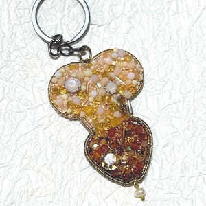 Könnte beinhalten: Ein Schlüsselanhänger mit Perlenbesatz, der aus zwei Teilen besteht: einem herzförmigen Boden und einer abgerundeten Oberseite. Der Schlüsselanhänger ist mit goldenen, orangefarbenen und cremefarbenen Perlen verziert und hat einen silberfarbenen Schlüsselring.