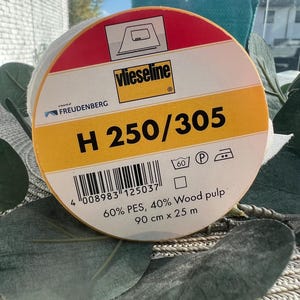 Könnte beinhalten: Eine Rolle Vlieseline H 250/305 Einlage. Das Etikett ist rot, weiß und gelb mit dem Markennamen und Produktdetails. Die Rolle ist 90 cm breit und 25 m lang, hergestellt aus 60 % PES und 40 % Holzstoff.