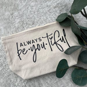 Könnte beinhalten: Eine cremefarbene Kosmetiktasche aus Canvas mit den Worten "ALWAYS be you tiful" in schwarzer Schrift. Die Tasche ist rechteckig mit Reißverschluss. Grüne Blätter sind im Hintergrund.