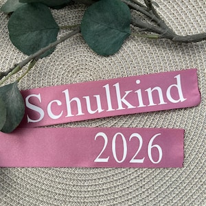 Könnte beinhalten: Zwei rosa Bänder mit weißem Text. Auf dem oberen Band steht "Schulkind" und auf dem unteren Band "2026". Grüne Blätter und Zweige sind im Hintergrund. Die Bänder liegen auf einer beigefarbenen, gewebten Oberfläche.