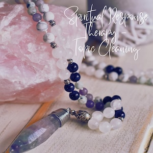 May include: A purple amethyst crystal pendant hangs from a beaded necklace with white, blue, and purple stones. The necklace is resting on a pink rose quartz stone. The text "Spiritual Response Therapy - Topic Cleaning" is written in white on a black background.