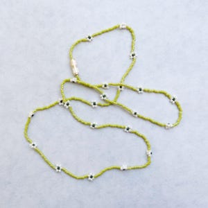 May include: A beaded necklace with a light green base and small white and black flower accents. The necklace has a magnetic clasp and is displayed on a light gray surface. The necklace is a delicate piece of jewelry.