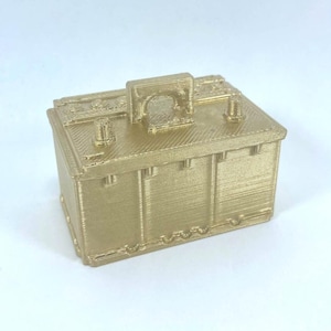 May include: A gold-colored, 3D-printed model of a car battery. The rectangular battery has detailed features, including terminals, a handle, and textured sides. The model is set against a plain white background.
