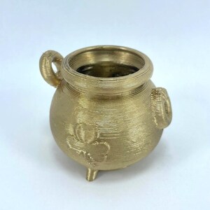 Può includere: Un piccolo calderone dorato con una superficie strutturata. Il calderone ha due manici e tre piedini. Un motivo a trifoglio è visibile sulla parte anteriore. Probabilmente utilizzato per scopi decorativi. Circa 8 cm di altezza.