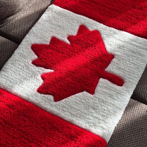Puede incluir: Un diseño rectangular de la bandera canadiense en rojo, blanco y rojo. La bandera presenta una hoja de arce roja en el centro sobre un fondo blanco. La bandera está hecha de un material suave y texturizado.