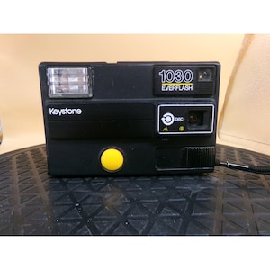 May include: Black Keystone 1030 Everflash camera with a yellow button and a flash. The camera has the word "Keystone" printed on the front. The camera has a black strap attached to the side.