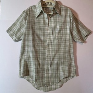 May include: A short-sleeved button-down shirt in a green and white plaid pattern. The shirt has a classic collar and is buttoned up. The shirt is hanging on a beige hanger.