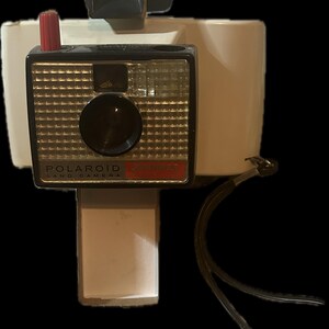 May include: A vintage white and black Polaroid Land Camera with a red button and a black strap. The camera has a silver lens and a black viewfinder.