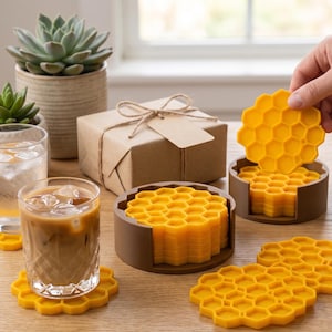 May include: Yellow honeycomb-shaped silicone coasters are displayed on a wooden table. The coasters are in a brown holder, and some are stacked. A hand is holding one coaster. There are two glasses with drinks, a gift box, and succulents.