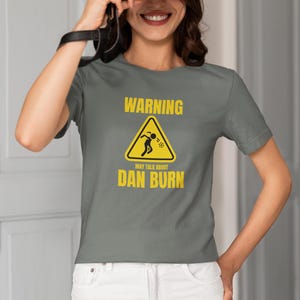 May include: A grey t-shirt with a yellow warning sign that says "WARNING MAY TALK ABOUT DAN BURN".