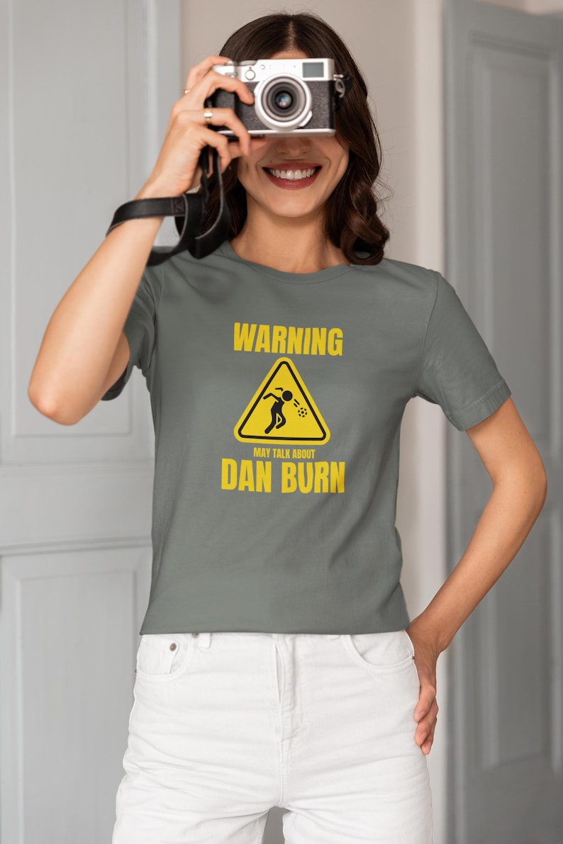 May include: A grey t-shirt with a yellow warning sign that says "WARNING MAY TALK ABOUT DAN BURN".