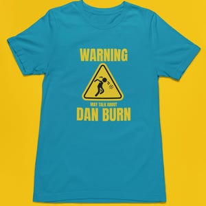 May include: A turquoise blue t-shirt featuring a yellow warning sign that reads "WARNING MAY TALK ABOUT DAN BURN". The sign depicts a stick figure vomiting.