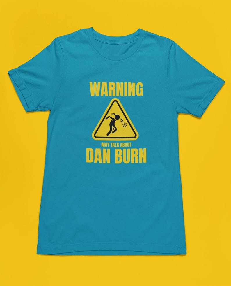 May include: A turquoise blue t-shirt featuring a yellow warning sign that reads "WARNING MAY TALK ABOUT DAN BURN". The sign depicts a stick figure vomiting.