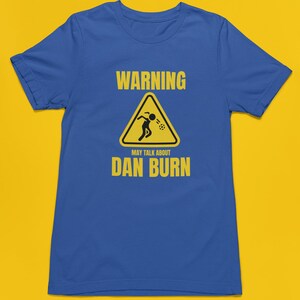 May include: A blue t-shirt featuring a yellow warning sign that reads "WARNING MAY TALK ABOUT DAN BURN". The sign depicts a stick figure vomiting.
