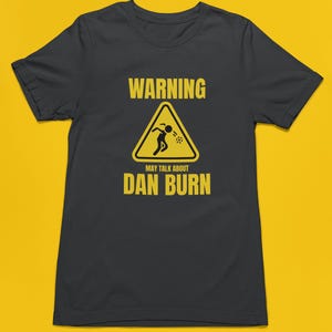 May include: A dark grey t-shirt with a yellow warning sign that says "WARNING MAY TALK ABOUT DAN BURN". The sign has a stick figure inside of it, throwing up.