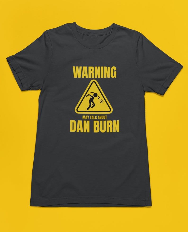 May include: A dark grey t-shirt with a yellow warning sign that says "WARNING MAY TALK ABOUT DAN BURN". The sign has a stick figure inside of it, throwing up.