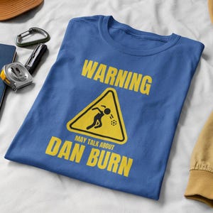 May include: A blue t-shirt featuring a yellow warning sign that reads "Warning May Talk About Dan Burn". The sign depicts a stick figure throwing a ball.