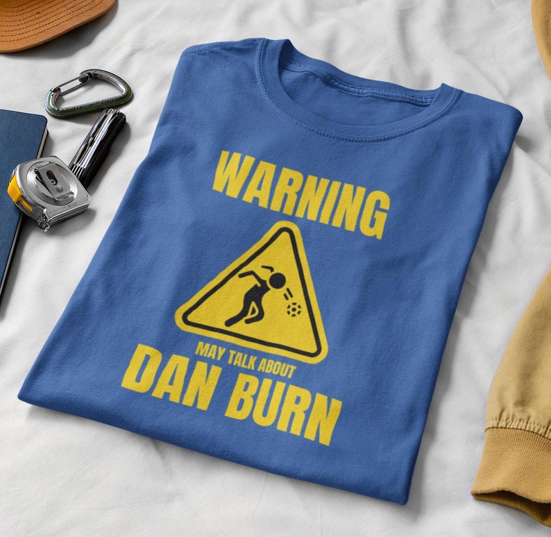 May include: A blue t-shirt featuring a yellow warning sign that reads "Warning May Talk About Dan Burn". The sign depicts a stick figure throwing a ball.
