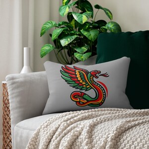 May include: A rectangular grey cushion with a colourful illustration of a winged serpent. The serpent design incorporates red, green, yellow, and black accents. The cushion is placed on a grey sofa with a cream-coloured blanket.