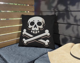 May include: Black decorative pillow featuring a white skull and crossbones design. The skull has a light gray outline and the bones are light gray with a white outline. The pillow is on a blue cushion.