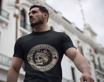 以下が含まれることがあります： 黒いTシャツ。円形のグラフィックには、王冠をかぶったスカルが描かれており、「PSYCHOBILLY ARTS MINNEAPOLIS」の文字が円形にデザインされています。デザインはタン色です。Tシャツは半袖です。