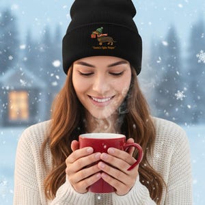 Könnte beinhalten: Eine schwarze Mütze mit einem gestickten Dackel, der einen Weihnachtshut trägt und ein Geschenk hält. Der Text "Santa's Little Helper" steht unter dem Dackel. Eine Person trägt die Mütze und hält eine rote Tasse.