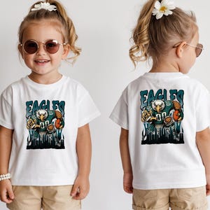 May include: White t-shirt with a cartoon eagle in a football uniform and the word "EAGLES". The eagle is holding a football. The shirt is worn by a young child.