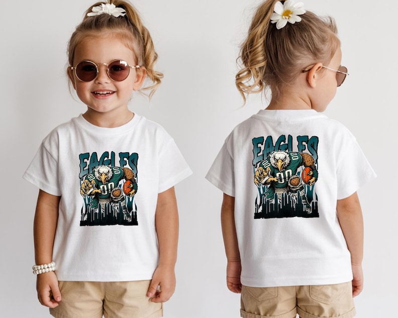 May include: White t-shirt with a cartoon eagle in a football uniform and the word "EAGLES". The eagle is holding a football. The shirt is worn by a young child.