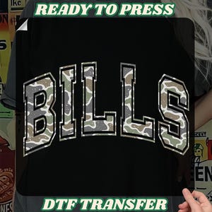 May include: Black t-shirt transfer with the word "BILLS" in a camouflage pattern, outlined in white. The text "READY TO PRESS" and "DTF TRANSFER" are displayed in green.