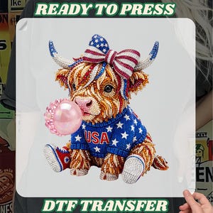 May include: DTF transfer featuring a Highland cow with a patriotic theme. The cow wears a blue sweater with white stars and the word "USA". It has a red, white, and blue bow and is blowing a pink bubble. The words "READY TO PRESS" and "DTF TRANSFER" are at the top and bottom.